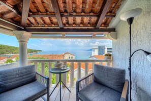 A balcony with two grey chairs, a small table with a vase and cup, and a lamp. The balcony overlooks a coastal view with buildings and a clear sky.