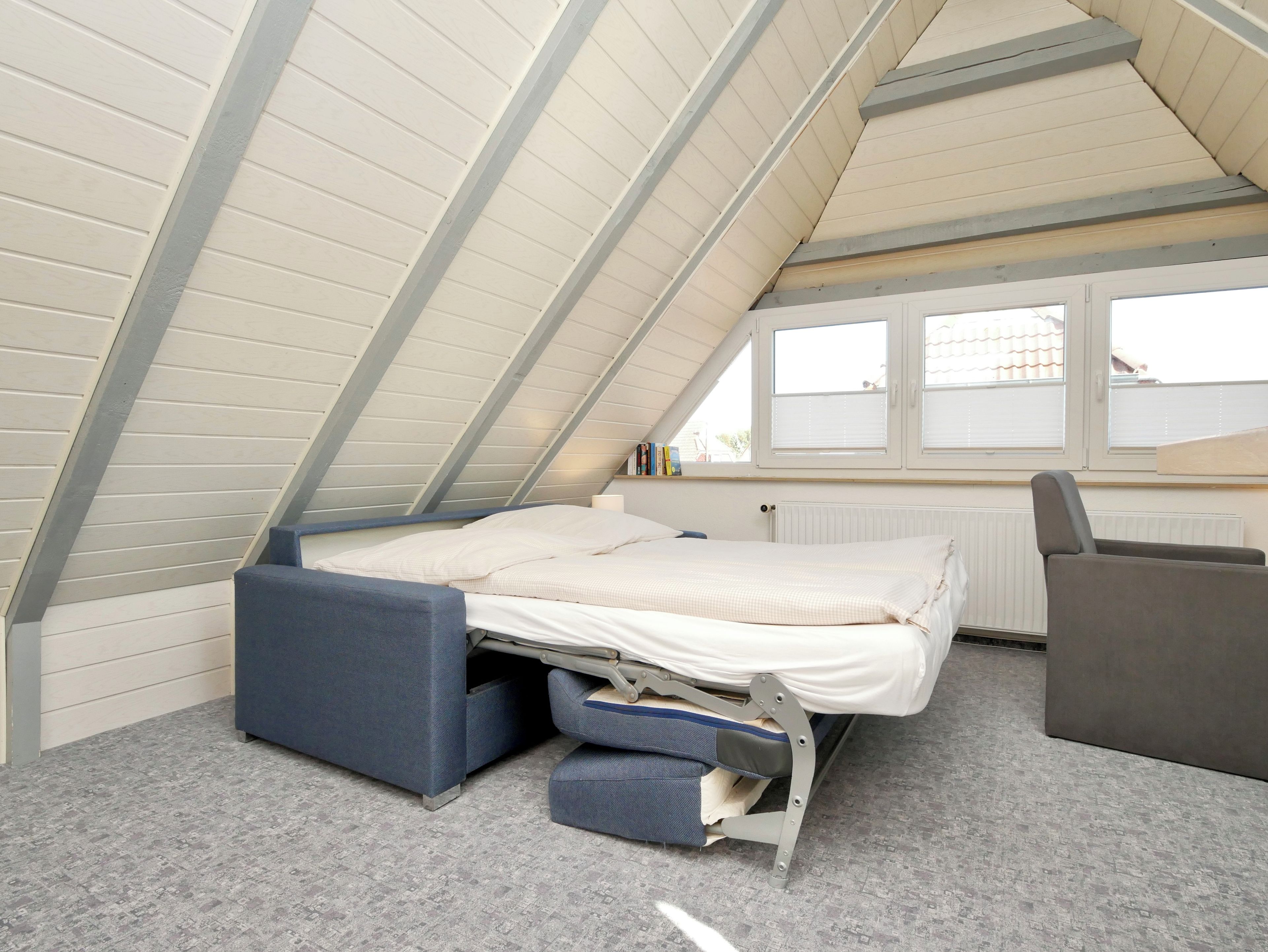 Ein kleiner Raum mit schrägem Dach, einem Bett, einem Sessel und einem Fenster.