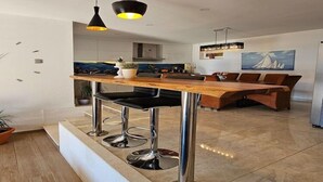 Une cuisine moderne dotée d’un îlot central en bois, de tabourets de bar, de luminaires suspendus et d’un coin repas offrant une vue sur l’océan.