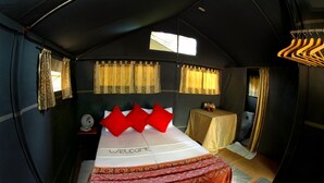 Una habitación tipo tienda de campaña con cama, una mesita con lámpara y una ventana con cortinas.