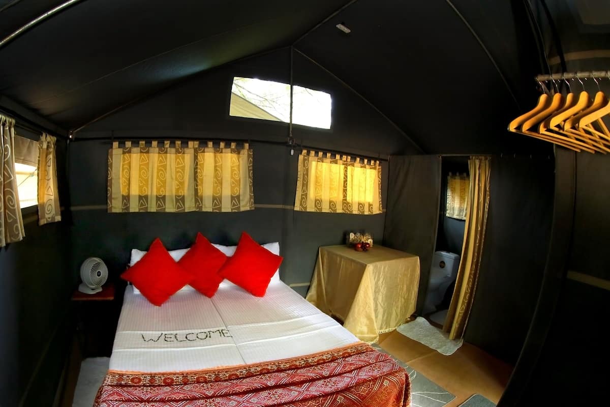 Una habitación tipo tienda de campaña con cama, una mesita con lámpara y una ventana con cortinas.