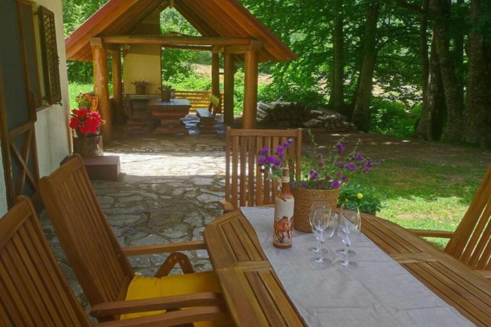 Een houten eethoek buiten, met stenen vloer, een gedekte tafel met wijnglazen en een fles, omringd door houten stoelen.