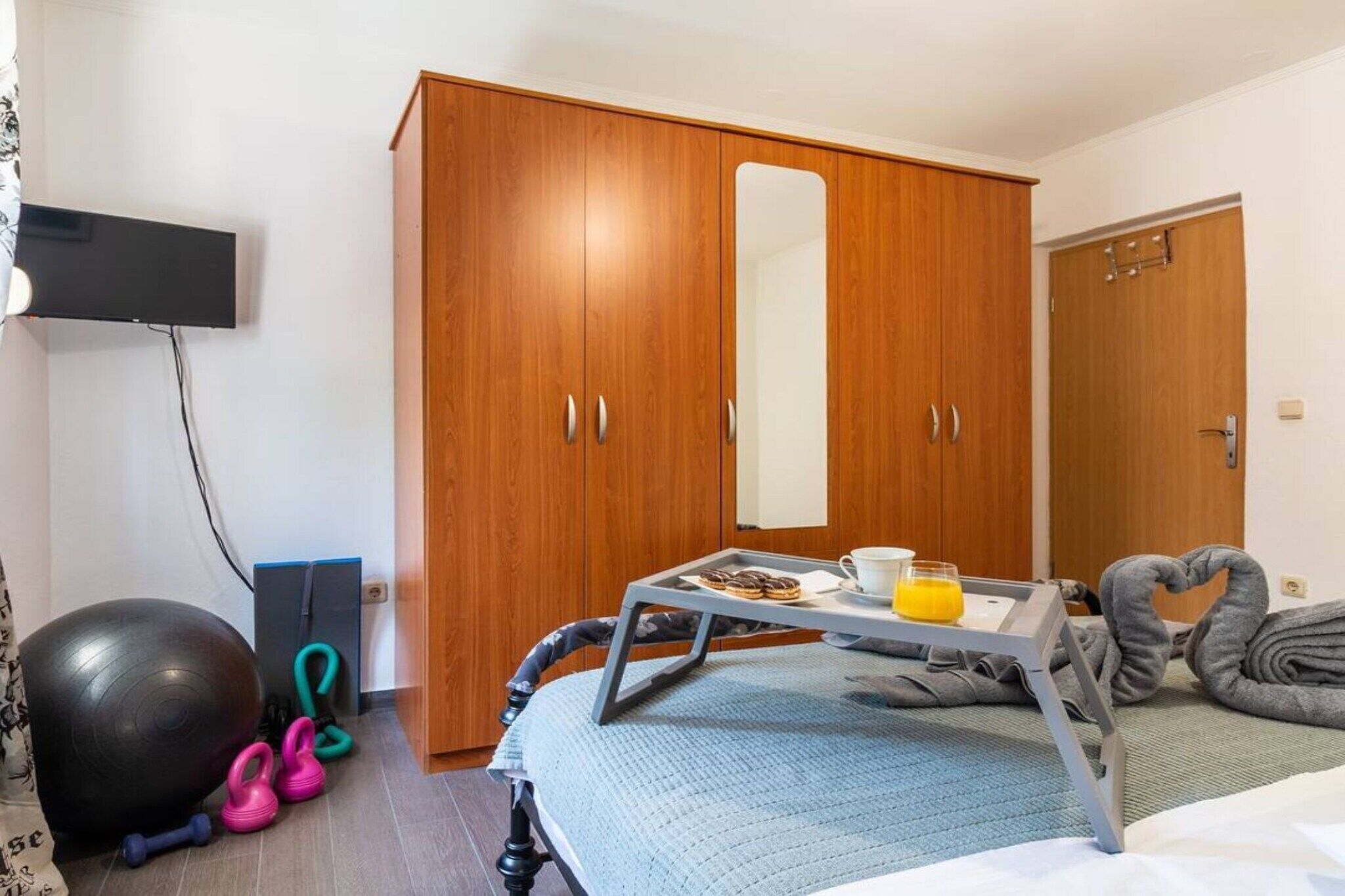 A bedroom with a bed, a wardrobe, a TV mounted on the wall, exercise equipment, and a tray with breakfast items.