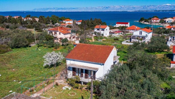A coastal village with white houses and red roofs, surrounded by greenery and a clear view of the sea and distant mountains.