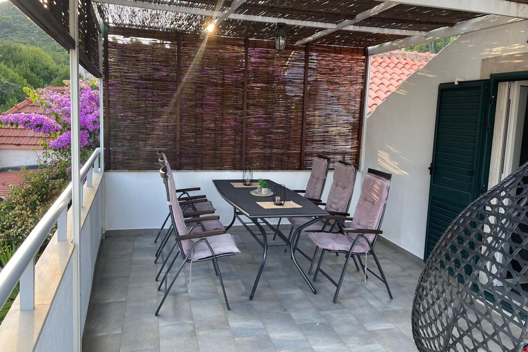 Un balcon avec une table et des chaises, un fauteuil en osier, une porte verte, et une vue sur des fleurs violettes et un toit recouvert de tuiles rouges.