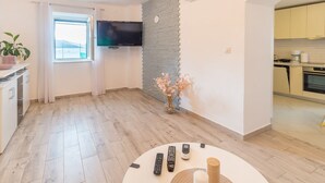 Una sala de estar moderna con un televisor de pantalla plana, una mesita con controles remotos, un ventanal con cortinas, una pared de ladrillo decorativa y una cocina visible a través de un arco.