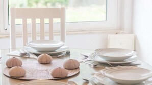 Mesa para comer con platos, tazones y cubiertos blancos, un camino de mesa tejido y conchas decorativas.