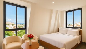 Quarto de hotel com cama, duas poltronas, uma mesinha redonda com um vaso de flores vermelhas e amplas janelas com vista para a cidade.