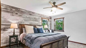 A bedroom with a wooden accent wall, a bed with a headboard, two bedside tables with lamps, a ceiling fan, and a window with a view of greenery. - Spacious Lake Arrowhead Golf Course Retreat (Nekoosa)