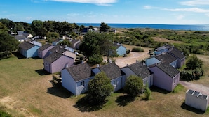 A cluster of small, single-story houses with sloped roofs, arranged in a semi-circular pattern around a central grassy area, with a view of the sea in the background. - Gite in Normandy Near Sandy Beach (Port-Bail-sur-Mer)