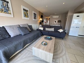 Un salon moderne avec un canapé gris, une table basse en bois et un coin cuisine doté d’un réfrigérateur blanc.