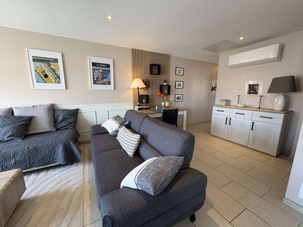 A modern living room with a grey sofa, a dining area with a table and chairs, and a kitchen with white cabinets.