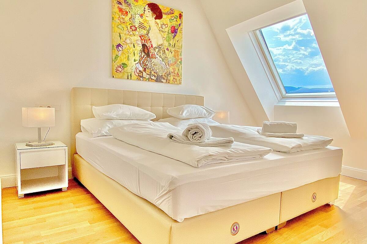 A bedroom with a bed, a nightstand, a painting, and a window with a view.