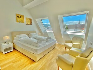 Ein Schlafzimmer mit einem Bett, einem Sessel, einem Nachttisch und zwei Fenstern mit Blick auf eine Kirche und die Stadt.