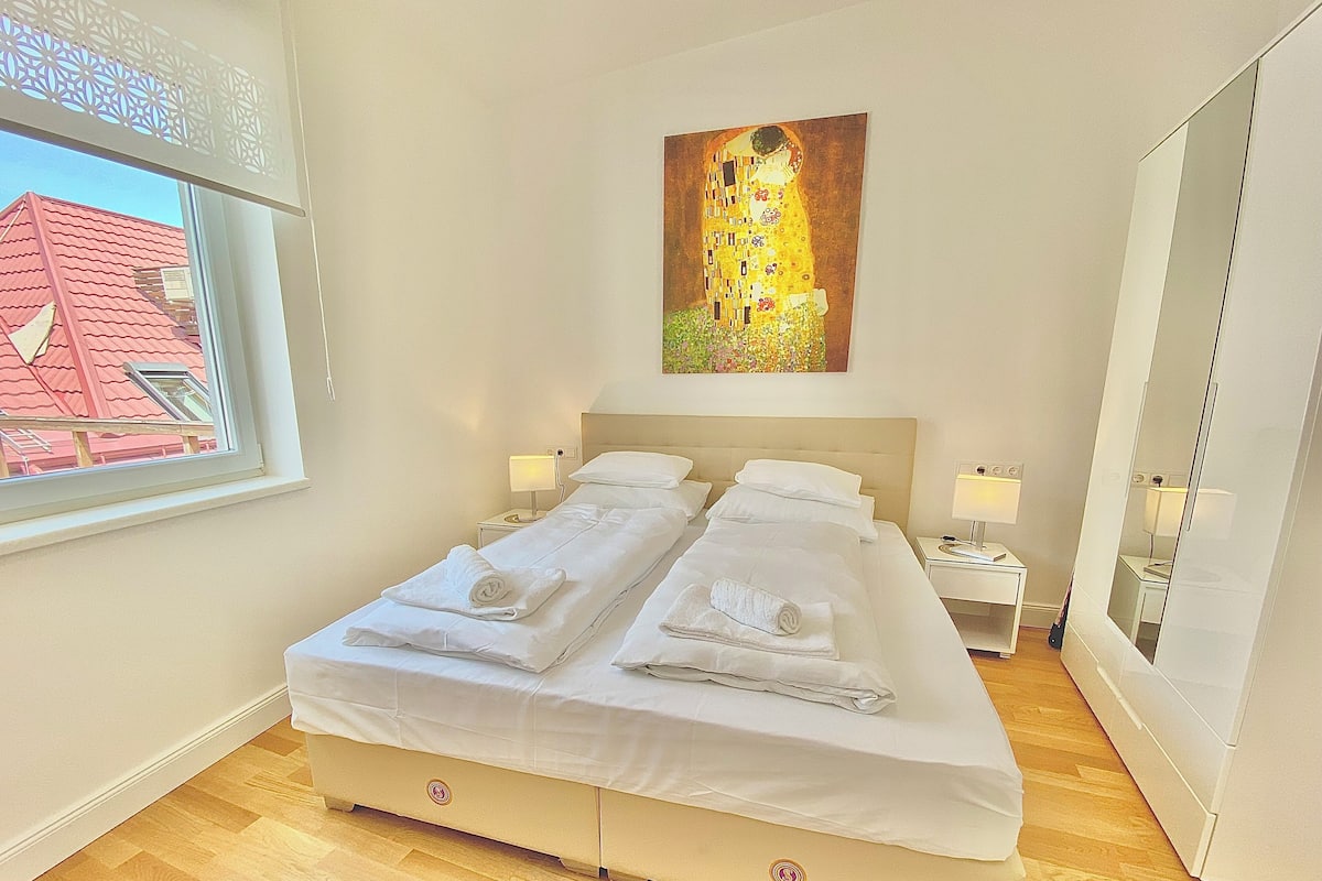 A hotel room with a large bed, two bedside tables, a window with a view of a red-roofed building, and a painting on the wall.