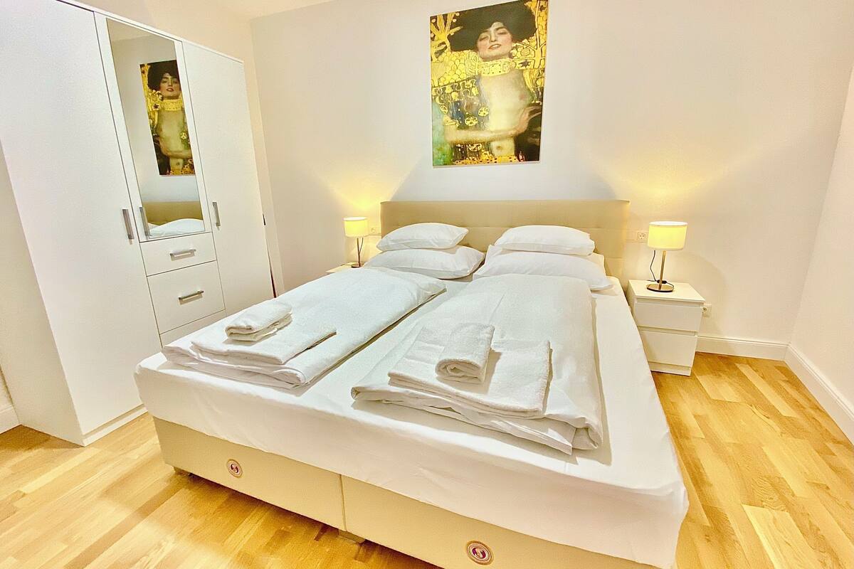A neatly made bed with white linens and towels, a white wardrobe, a bedside table with a lamp, and a framed artwork on the wall.