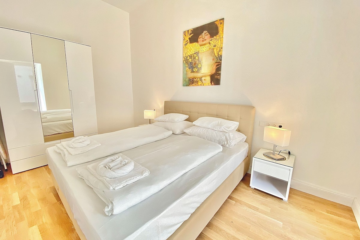 A neatly made bed with white linens and towels, a bedside table with a lamp, a large mirror, and a painting on the wall.