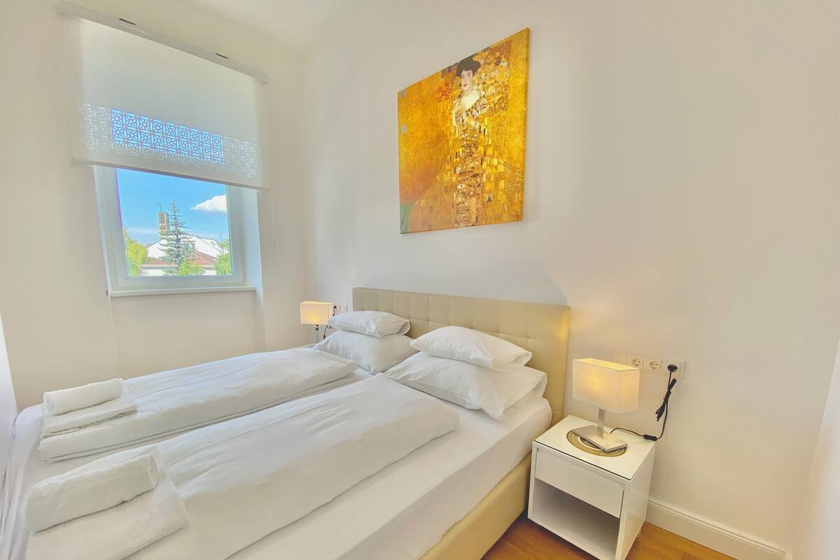 A hotel room with a bed, two bedside tables, a window with a view, and a painting on the wall.