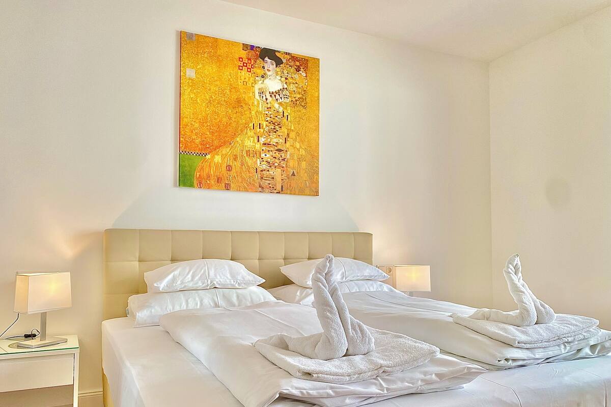 A hotel room with a large bed, white linens, two bedside lamps, and a painting on the wall.