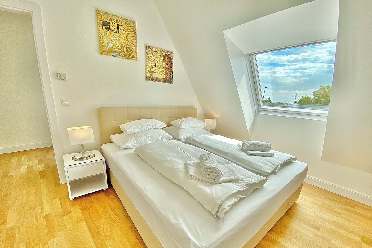 A modern bedroom with a large bed, a nightstand, a window with a view, and two wall art pieces.