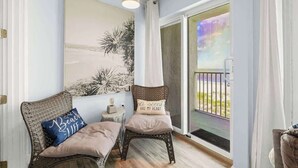 Une pièce comprenant un fauteuil en osier, un banc rembourré, une œuvre d’art accrochée au mur et une porte-fenêtre coulissante donnant sur un balcon.