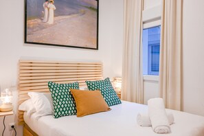 Un lit bien fait, avec des draps blancs, deux coussins à motifs et un coussin décoratif marron. Un tableau représentant deux personnages est accroché au mur, au-dessus du lit.