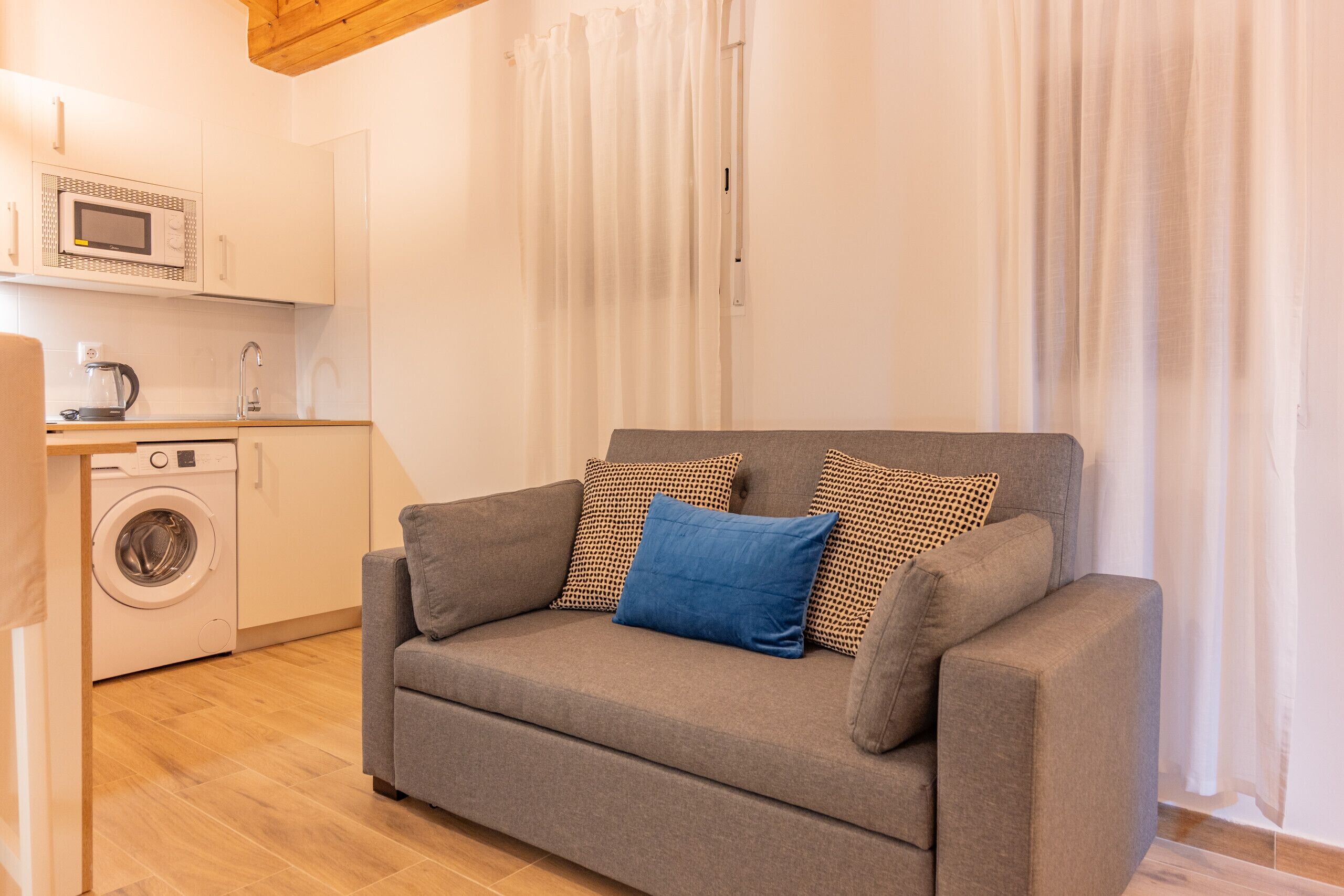 Un espace de vie compact comprenant un canapé gris, un lave-linge et un four à micro-ondes dans un coin cuisine.