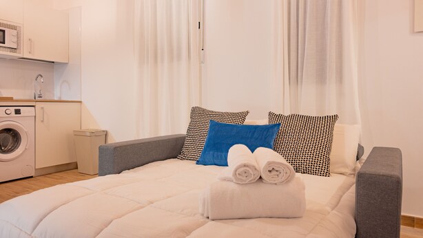 A neatly made bed with rolled towels, a washing machine, and a small trash bin in a clean, well-lit room with white walls and curtains.
