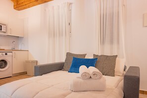 A neatly made bed with rolled towels, a washing machine, and a small trash bin in a clean, well-lit room with white walls and curtains.