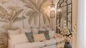 Una sala de estar con un sofá, almohadones decorativos, un jarrón con flores y una pared con un mural tropical.