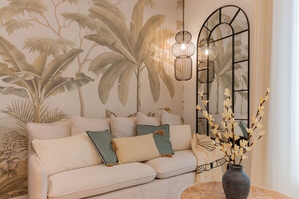 Una sala de estar con un sofá, almohadones decorativos, un jarrón con flores y una pared con un mural tropical.
