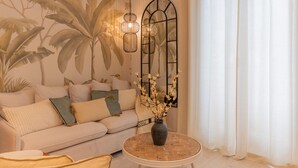 Una sala de estar con un sofá, una mesa redonda de centro, un jarrón con flores, un mural en la pared y una ventana con cortinas.