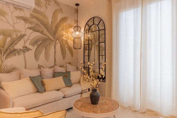 Una sala de estar con un sofá, una mesa redonda de centro, un jarrón con flores, un mural en la pared y una ventana con cortinas.