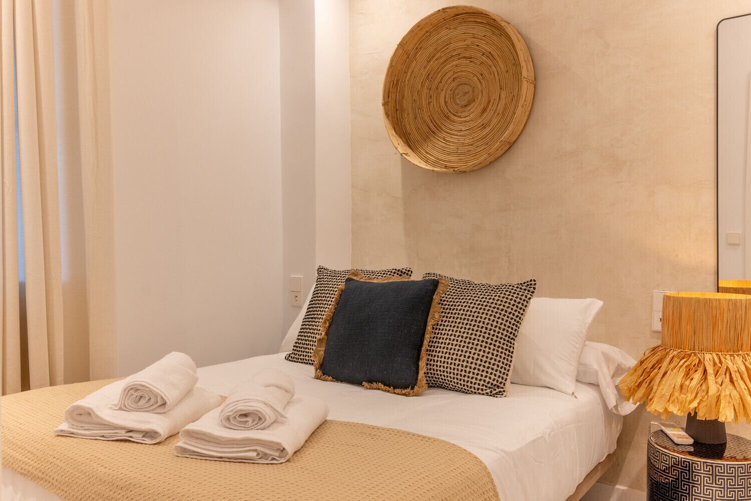 A neatly made bed with rolled towels, a round woven wall hanging, and patterned pillows.