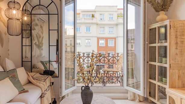 Un balcón con vistas a edificios, un sofá con cojines, un jarrón con ramas secas y un armario con puertas de cristal.