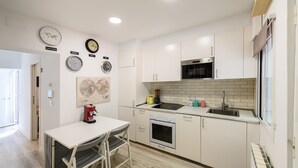 A modern kitchen with white cabinets, a microwave, an oven, and a sink. There are clocks on the wall and a table with chairs.