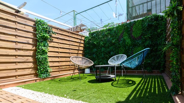 A patio with a wooden fence, two white chairs, a small table, and a green wall with plants.