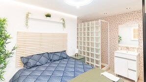 A bedroom with a bed, a bedside table, a nightstand, a wall-mounted shelf with plants, a window with blinds, and a bathroom with a sink and mirror.