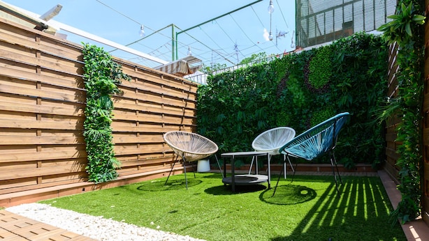 Un patio con cerca de madera, dos sillas blancas, una mesa pequeña y una pared verde con plantas.