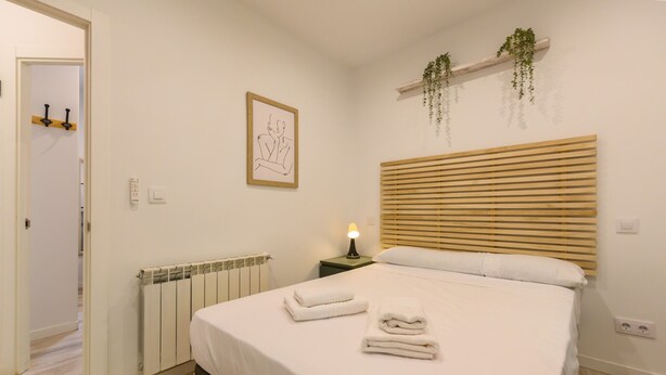 Een netjes opgemaakt bed met witte lakens en handdoeken, een bedlampje, een radiator, een lijst met een kunstwerk aan de muur, en een houten hoofdbord.