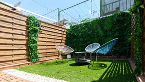 Een terras met een houten omheining, twee witte stoelen, een kleine tafel en een groene wand met planten.