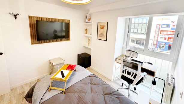 Une chambre à coucher moderne, équipée d’un lit, d’une table de chevet, d’un téléviseur et d’une fenêtre donnant sur des immeubles.