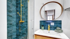 Ein Badezimmer mit Duschbereich, blauem Fliesenbelag, einem weißen Waschbecken mit Goldarmatur und einem Holz-Waschtisch.
