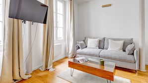 Ein modernes Wohnzimmer mit einem flachbildfernseher an der Wand, einem grauen Sofa mit Kissen, einem Holztisch und einem großen Fenster mit Vorhängen.