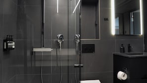 Uma casa de banho moderna com cabine de duche em vidro, lavatório com torneira e suporte para rolo de papel higiénico.