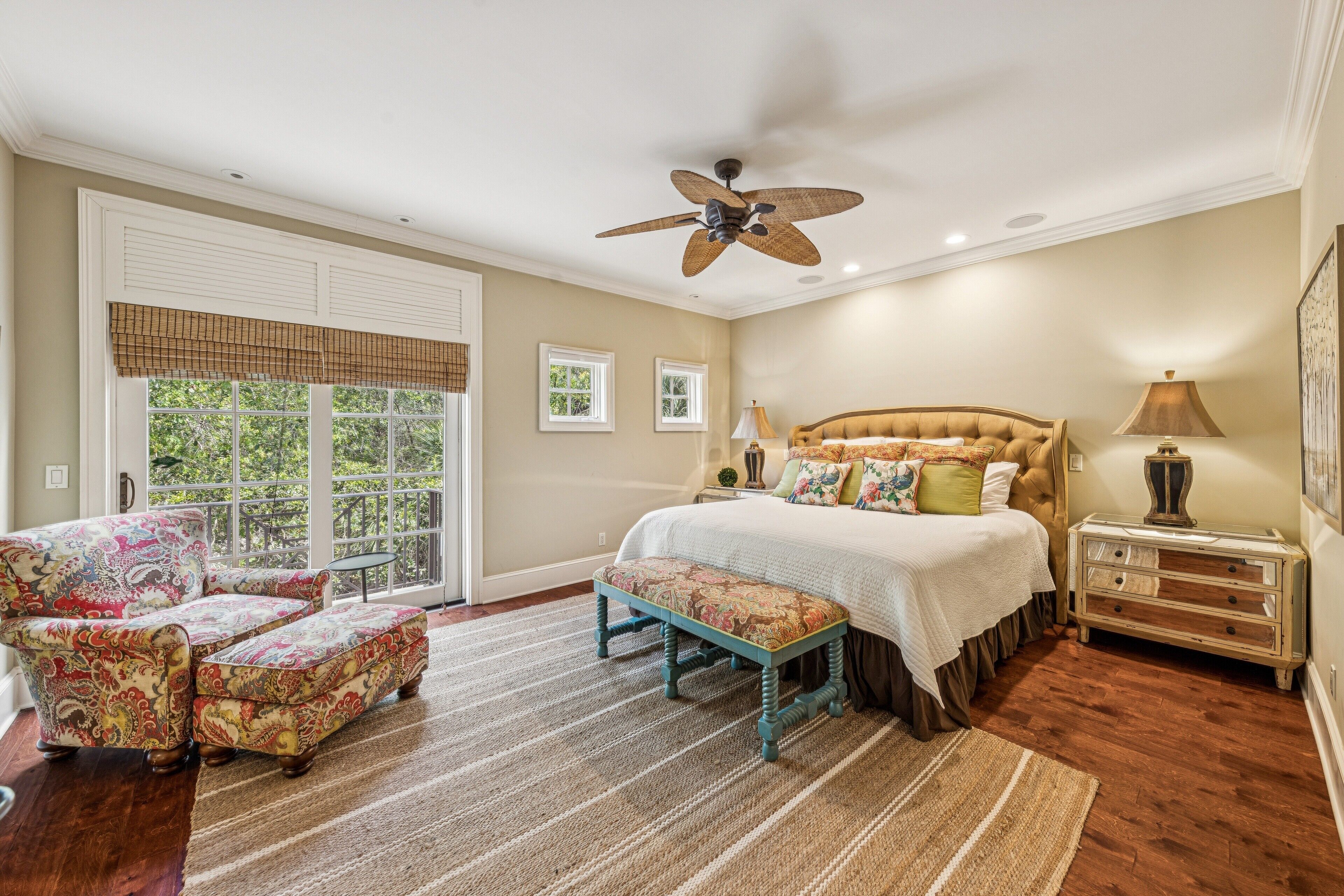 A bedroom with a large bed, a ceiling fan, a window with blinds, and a patterned armchair.