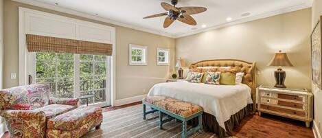 A bedroom with a large bed, a ceiling fan, a window with blinds, and a patterned armchair.