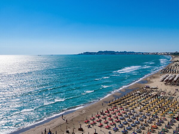 A beach with rows of umbrellas and sun loungers, a clear blue sea, and a distant city skyline.