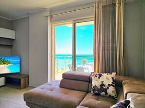 A modern living room with a large flat-screen TV, a sofa, and a view of the ocean through a sliding glass door.
