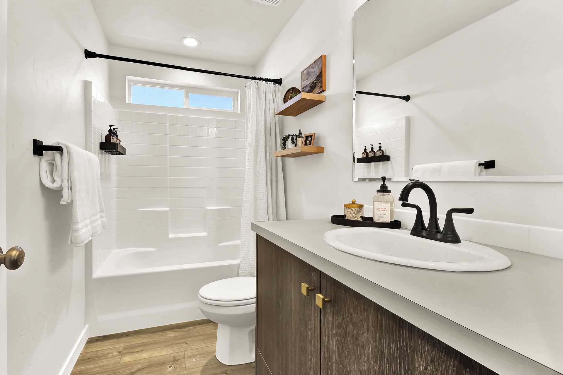 A modern bathroom with a white sink, black faucet, and a wooden vanity. There is a shower curtain, a toilet, and a window above the shower.